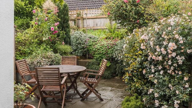 A patio with a round table and four chairs, surrounded by lush greenery and flowering plants.