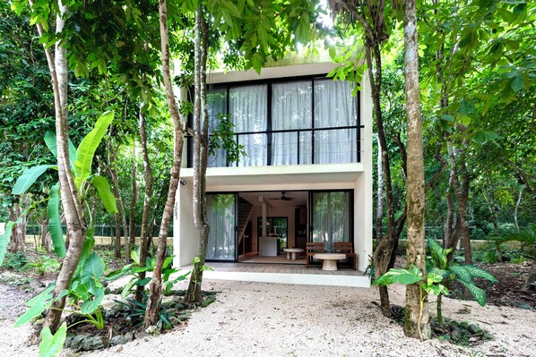 A modern two-story house with a large glass door entrance, surrounded by lush greenery.