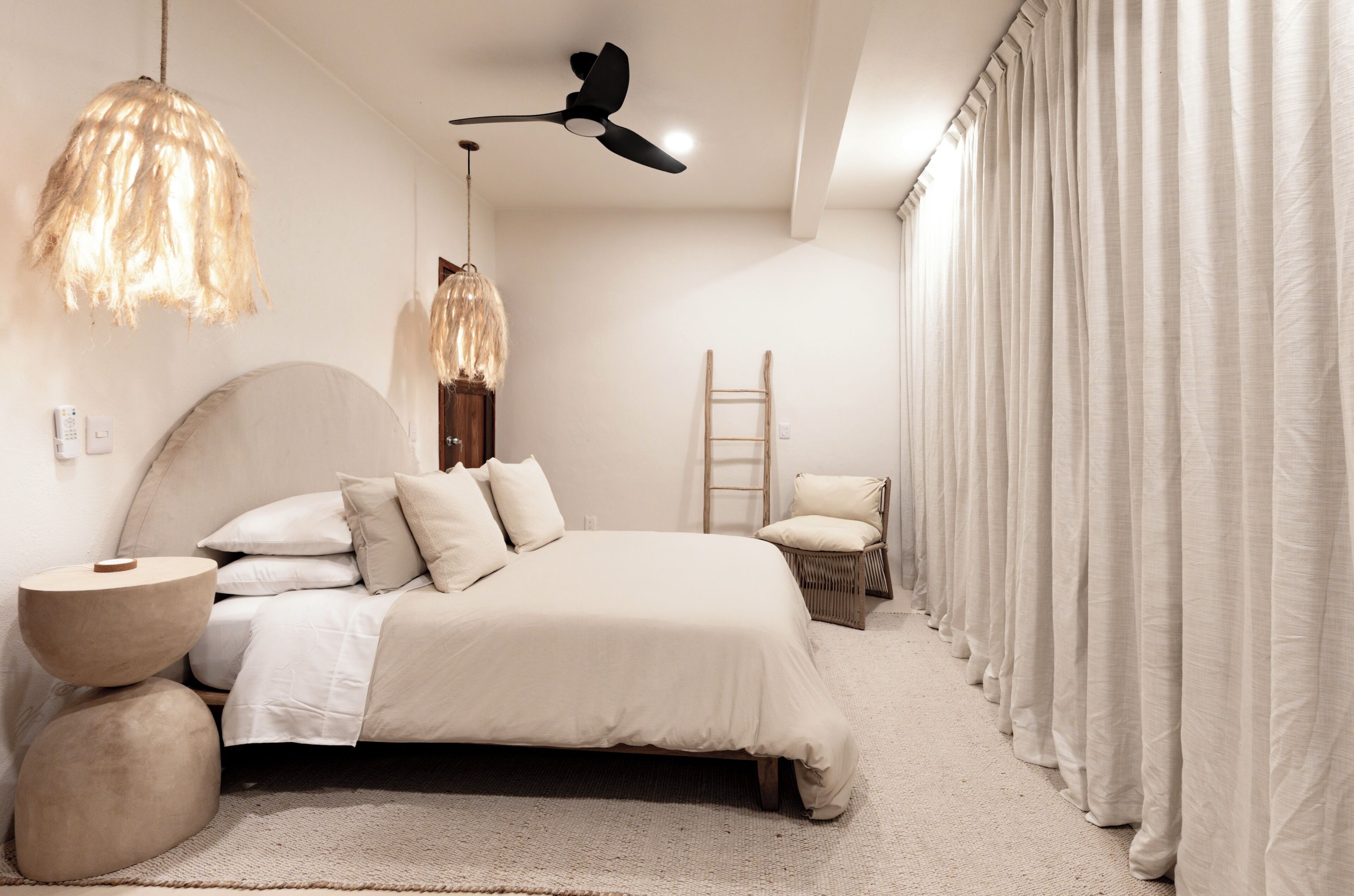 Une chambre à coucher comprenant un lit, un ventilateur de plafond, un luminaire suspendu, une chaise et une fenêtre avec des rideaux.