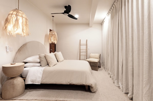 Ein Schlafzimmer mit einem Bett, einem Deckenventilator, einer Hängelampe, einem Sessel und einem Fenster mit Vorhängen.