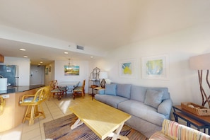 A modern living room with a sofa, a dining table, and chairs. There is a kitchen area with a refrigerator and a microwave.
