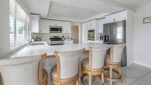 A modern kitchen with white cabinetry, a stainless steel refrigerator, a microwave, and a gas stove. There are three bar stools with cushioned seats and wooden legs.