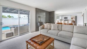 A modern living room with a large grey sofa, a wooden coffee table, and a view of a swimming pool through a sliding glass door.