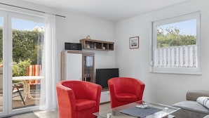 Un salon moderne avec une table basse en verre, deux fauteuils rouges, un canapé gris et un téléviseur posé sur un meuble en bois.
