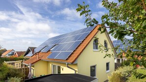 Une maison avec un toit en pente recouvert de panneaux solaires, entourée de verdure et d’autres maisons.