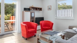 A modern living room with a glass coffee table, two red armchairs, a grey sofa, and a TV on a wooden stand. - Group house with sauna on the Baltic Sea (Boiensdorf)