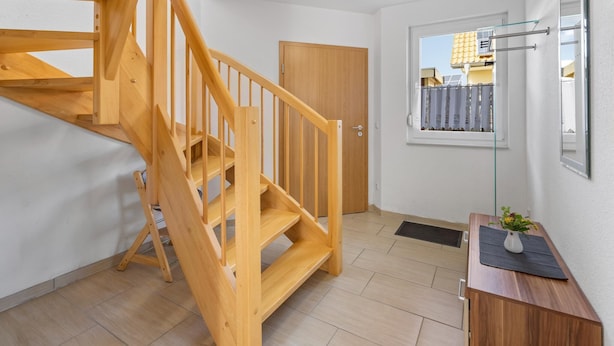 A wooden staircase leading to a second floor, a door, a window with a view of buildings, and a small table with a vase. - Group house with sauna on the Baltic Sea (Boiensdorf)