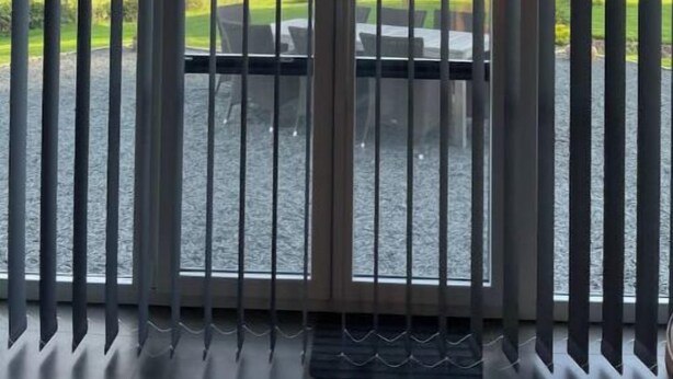 A room with a large window featuring vertical blinds, a view of a grassy area with a table and chairs outside.