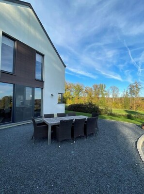 A modern building with a gravel patio, a dining table with chairs, and a view of trees and grass.