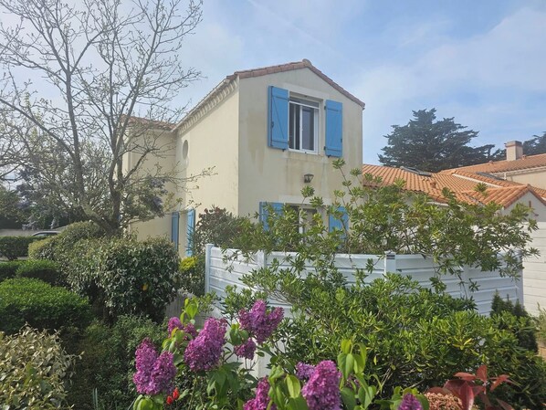 A two-story house with blue shutters, surrounded by lush greenery and purple flowers. - Marine air (Barbâtre)