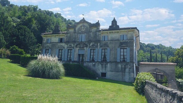 A large, historic building with multiple windows and a central entrance, surrounded by lush greenery and a hilly landscape.