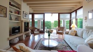A spacious living room with a white sofa, two wicker chairs, a coffee table, and a flat-screen TV. Large windows offer a view of greenery outside.