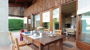 A wooden dining table with chairs, a glass door leading to a kitchen, and a view of a garden.