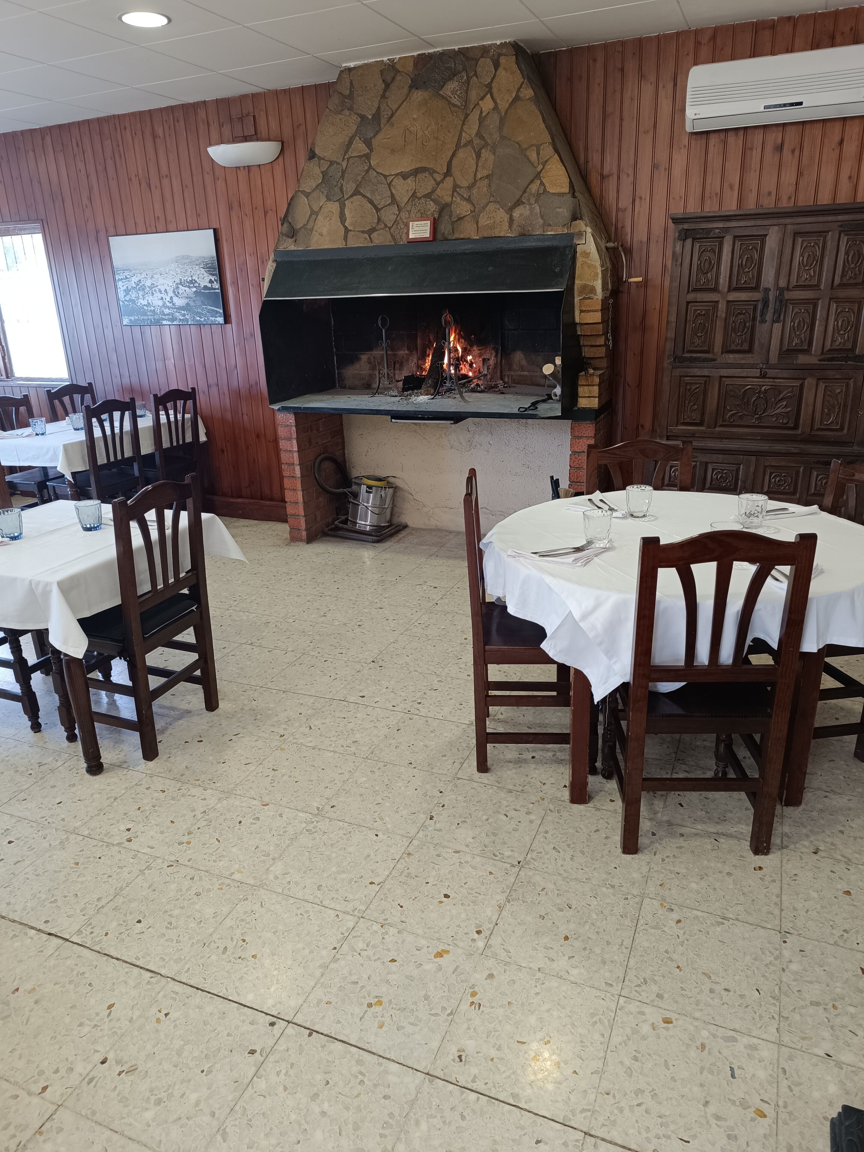 A restaurant with a stone fireplace, wooden walls, and tiled flooring. Tables and chairs are set for dining.