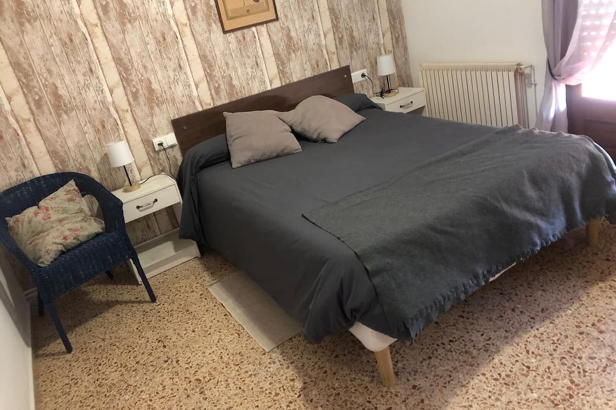 Une chambre avec un mur en bois, un lit recouvert d’une couverture grise, deux tables de chevet avec des lampes, une chaise avec un coussin à motifs floraux et un radiateur.