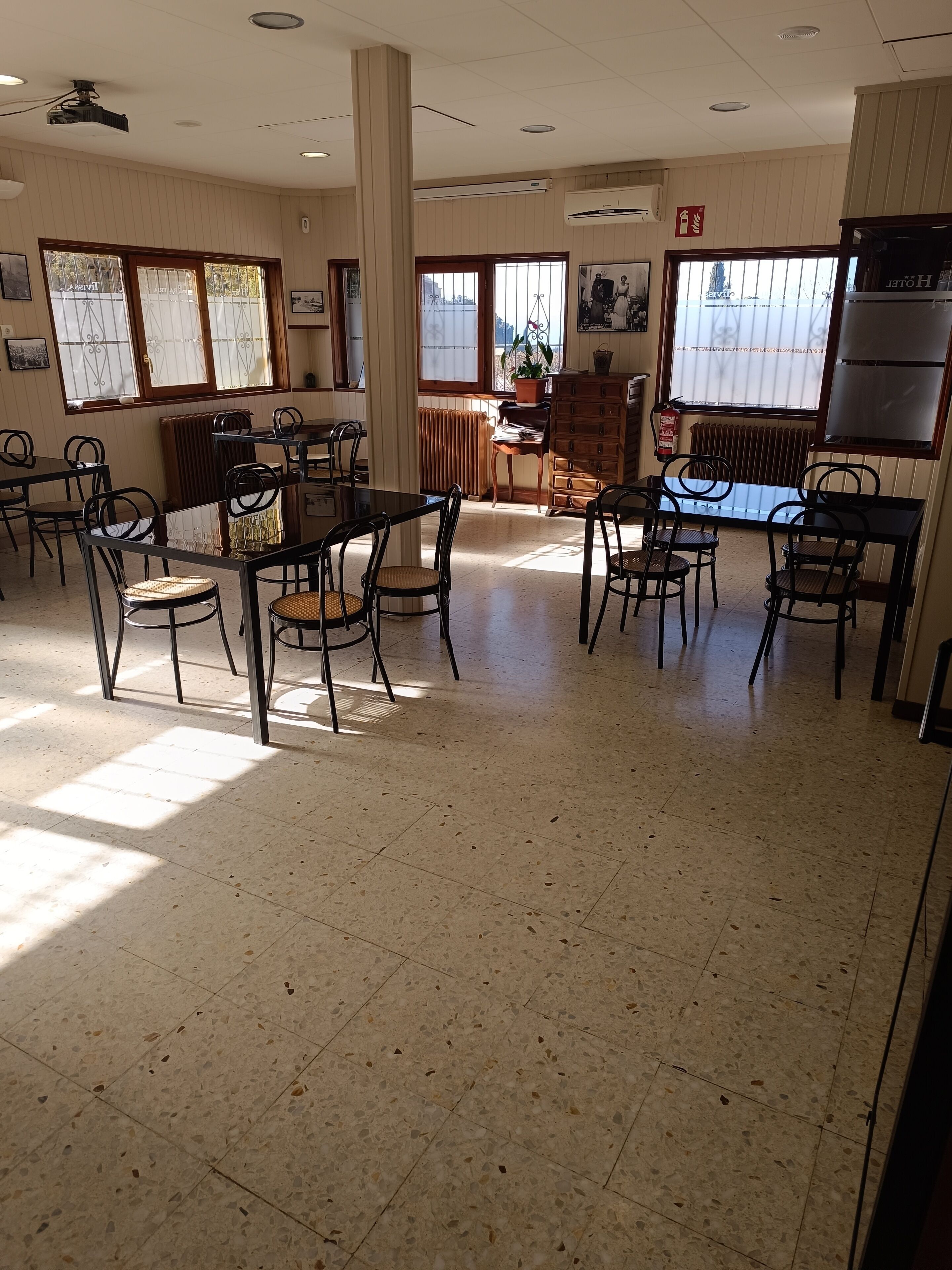 Un vaste espace de restauration dans l’hôtel, avec de nombreuses tables et chaises, de grandes fenêtres équipées de stores et un extincteur fixé au mur.