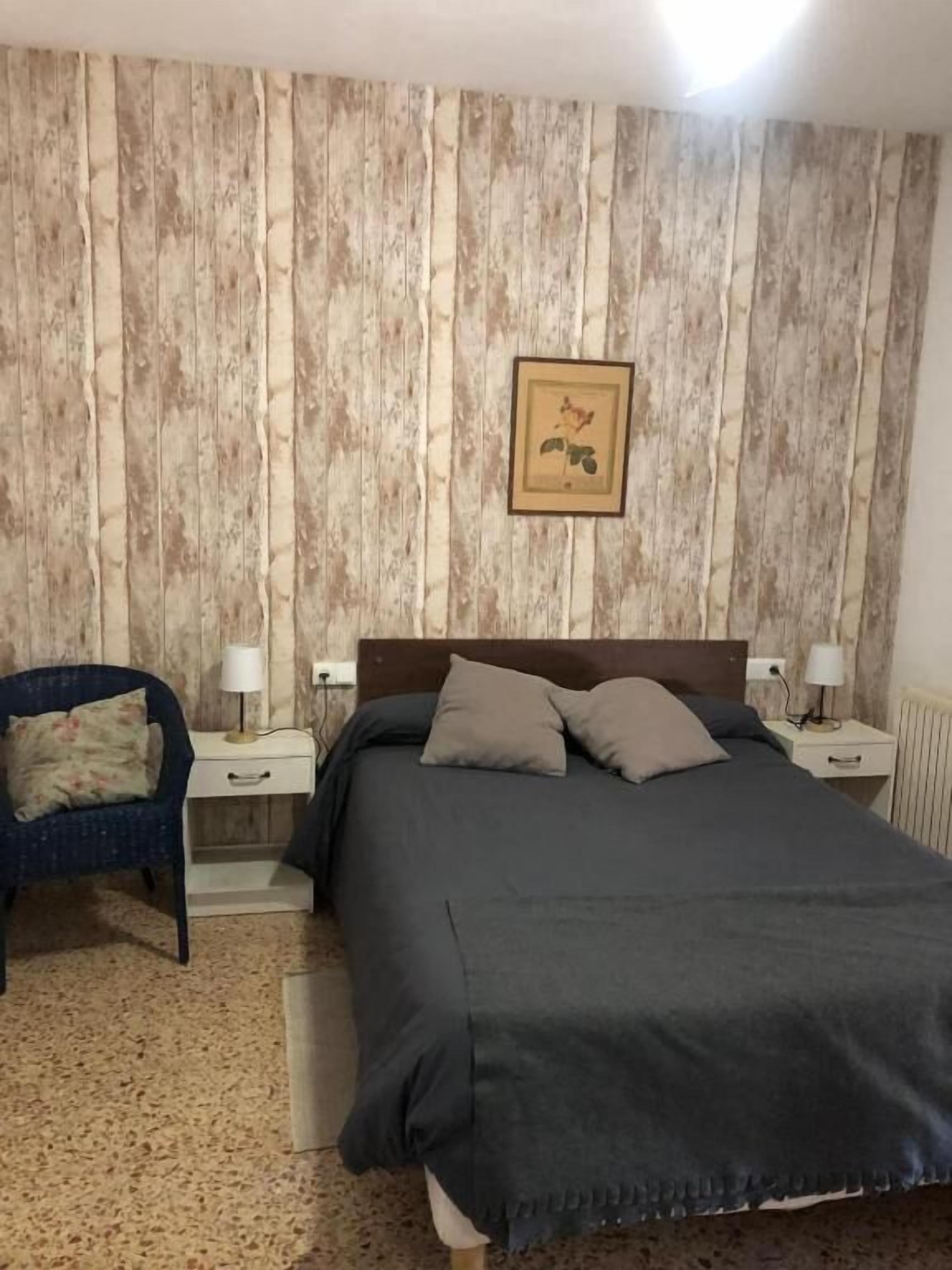 Ein Schlafzimmer mit Holzpaneelen, einem Bett mit grauer Bettwäsche, zwei Nachttischen mit Lampen, einem Sessel und einem gerahmten Bild an der Wand.