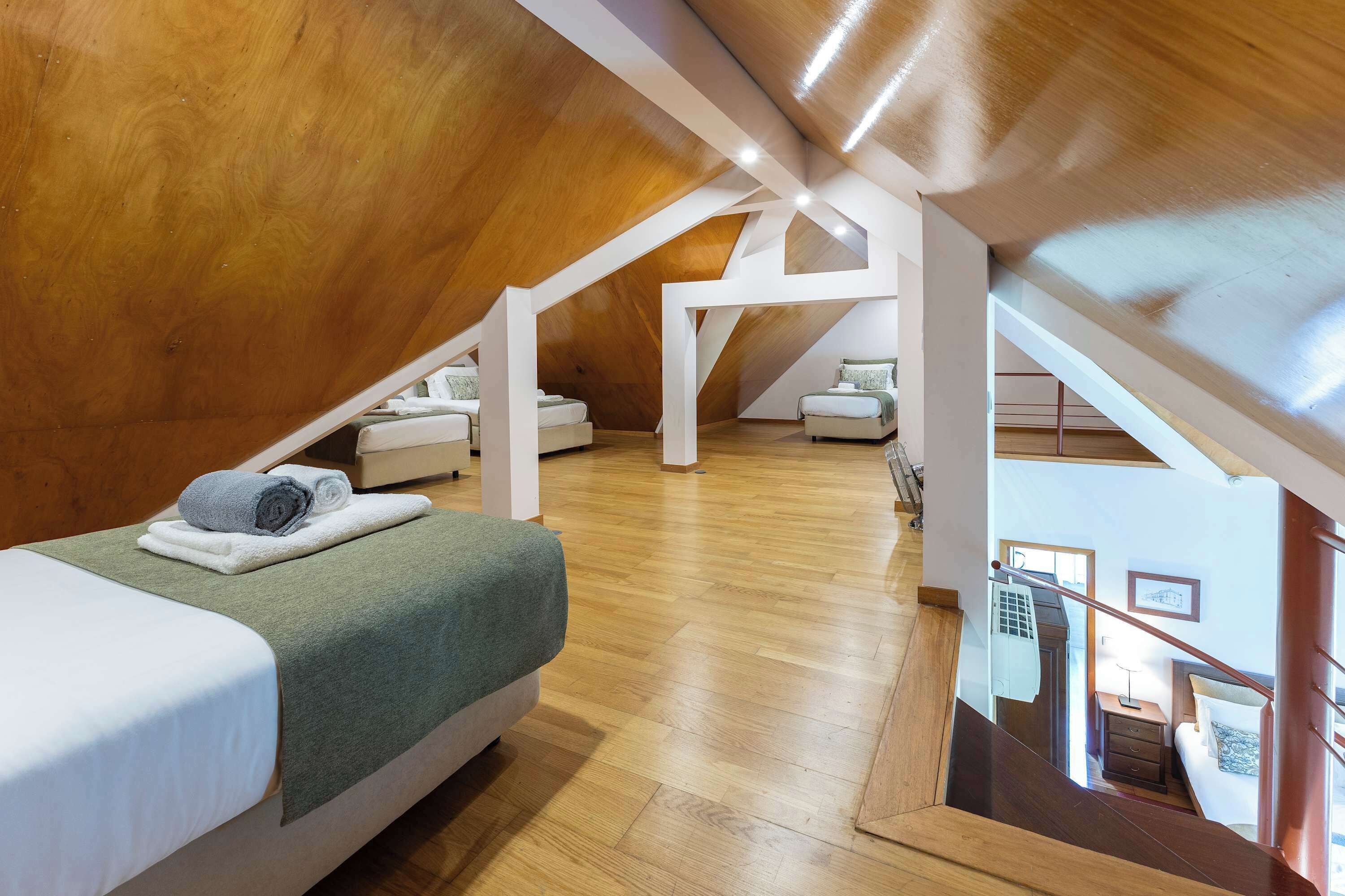 A bedroom with a sloped ceiling, wooden flooring, a bed with a green blanket, and a view of the adjacent room through a glass door.