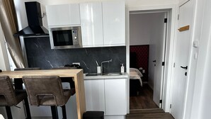 Una sala moderna con un sofá, una mesa de centro con un florero y un control remoto, una cocina con alacenas blancas y un microondas, y un pasillo que conduce a un dormitorio.