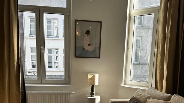 Una sala de estar con un sofá, una mesa redonda de centro con un jarrón y un control remoto, una lámpara y un cuadro en la pared.