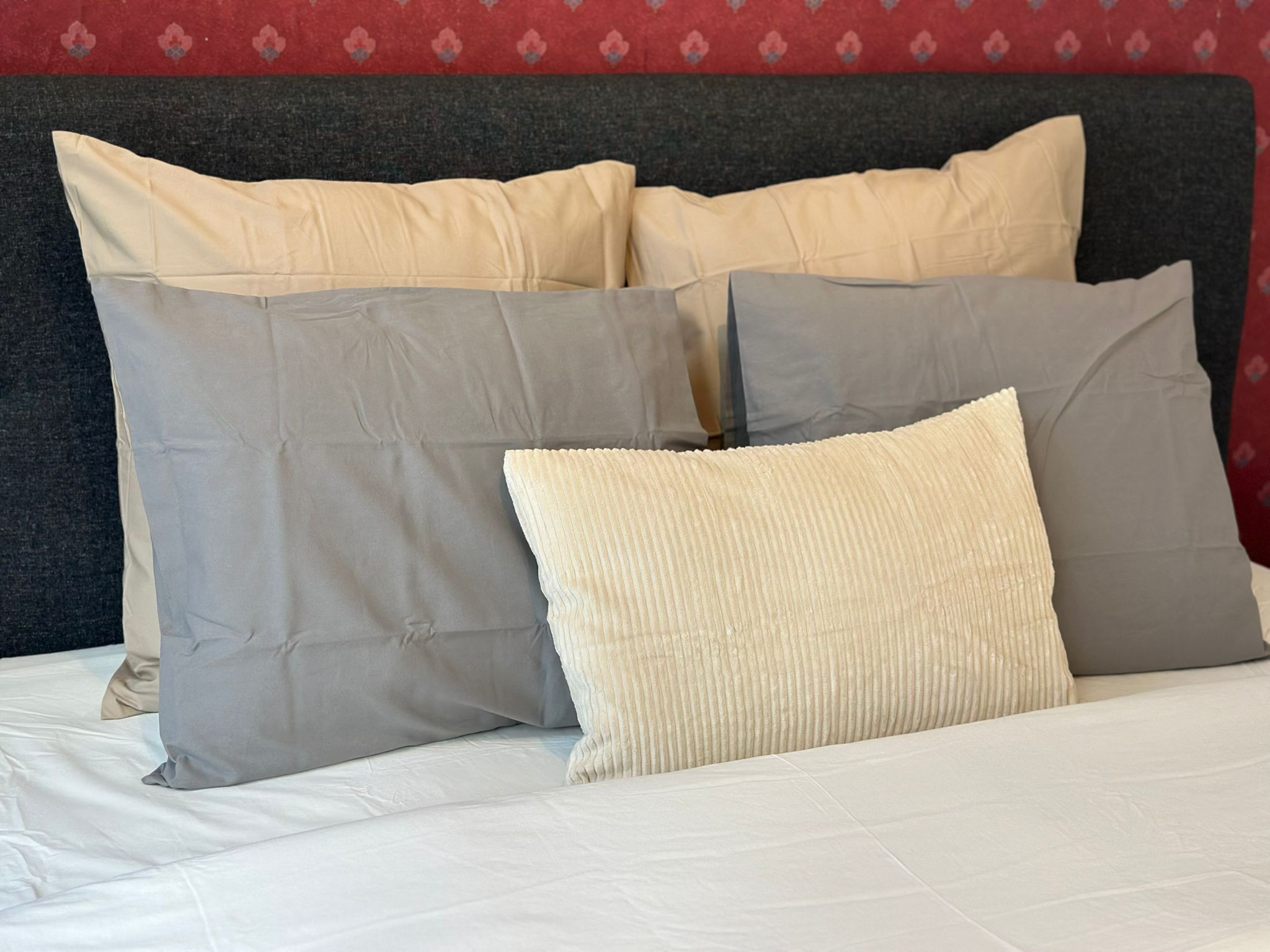 Una cama con varios almohadones en tonos beige y gris, un cabecero oscuro y una pared con estampado al fondo.