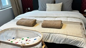 Una cama bien hecha con sábanas blancas y dos toallas dobladas, una cuna de bebé con malla blanca y diseños coloridos, y un cabecero rojo estampado.