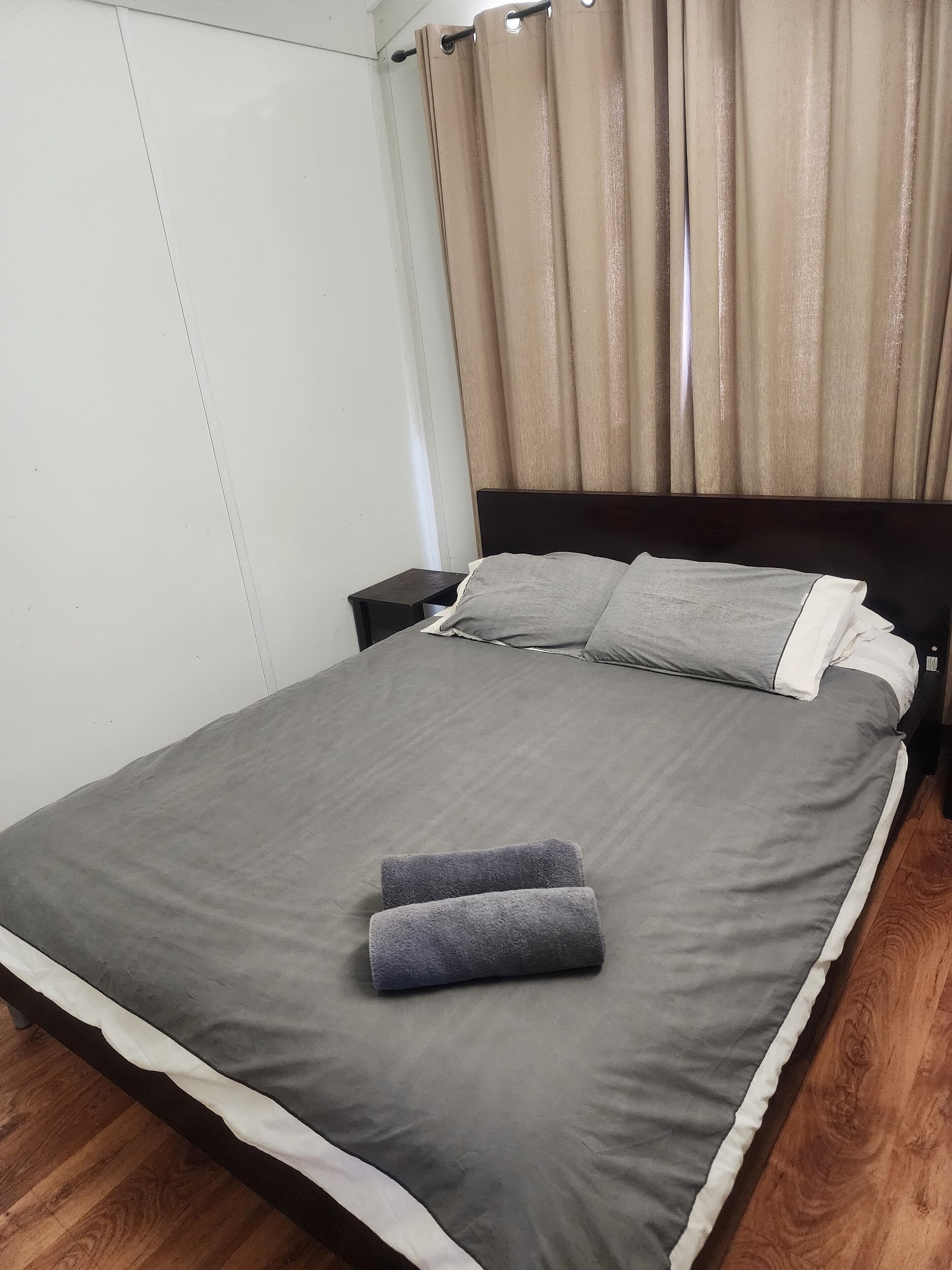 Cama bem arrumada com colcha cinza e almofadas, mesa de cabeceira de madeira com um telefone, e cortina cobrindo uma janela.
