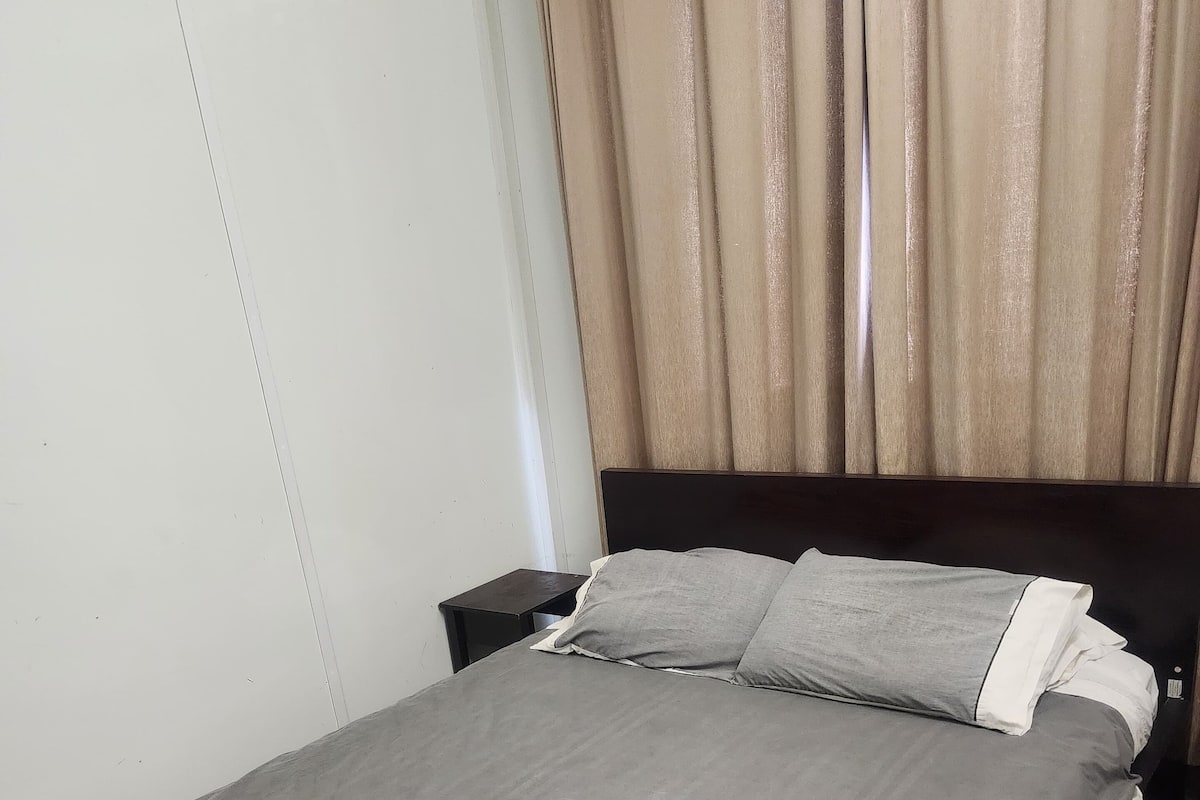 A neatly made bed with a grey bedspread and pillows, a wooden nightstand with a phone, and a curtain covering a window.