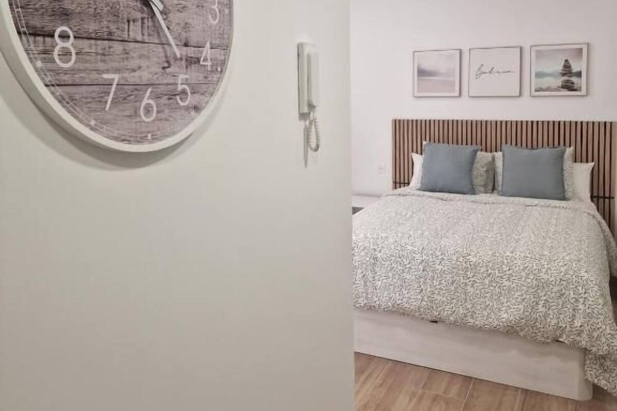 Une chambre à coucher avec une horloge en bois accrochée au mur, un lit recouvert d’une couvre-lit à motifs et une table de chevet avec une lampe.