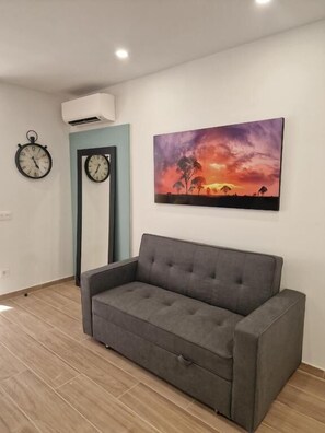 Olohuone, jossa on harmaa sohva, kaksi seinäkelloa ja auringonlaskua kuvaava taulu.