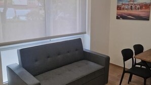 Un soggiorno moderno con un divano grigio, un tavolo da pranzo con sedie e un'ampia finestra con vista su un edificio.