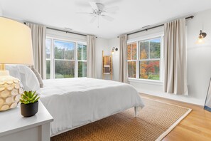 A bedroom with a large bed, a ceiling fan, a window with curtains, a bedside table with a lamp, and a view of autumn trees. - Spacious Living, Covered Porch & Forest Setting | Beaver Creek by AvantStay (Stockbridge)