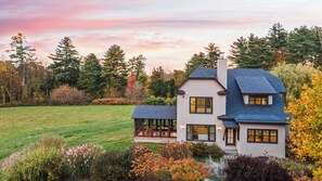 A two-story house with a large lawn, a patio with outdoor furniture, and a view of the surrounding trees and sky. - Spacious Living, Covered Porch & Forest Setting | Beaver Creek by AvantStay (Stockbridge)