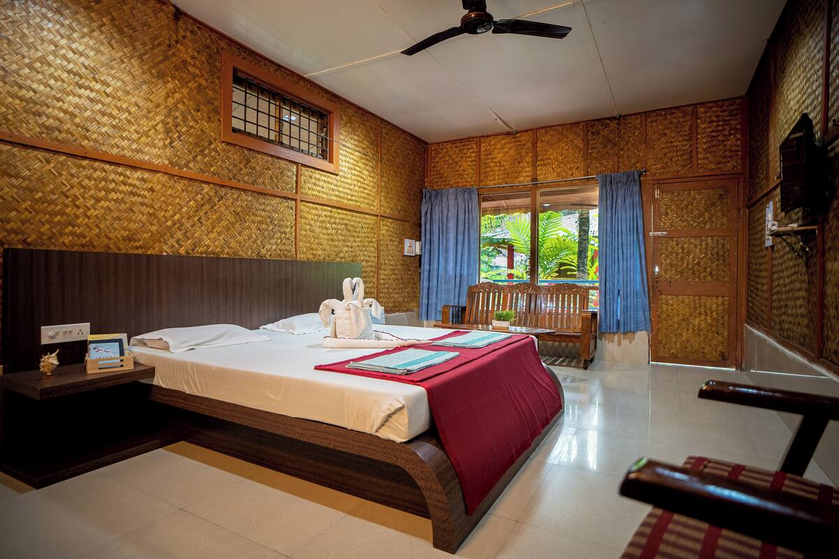 A hotel room with a large bed, wooden walls, a ceiling fan, and a view of greenery outside.