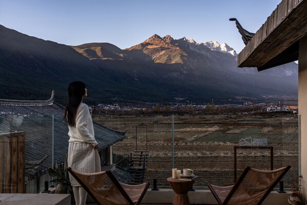Una mujer de pie en un balcón con vistas a un paisaje montañoso que incluye un valle y edificios al fondo.