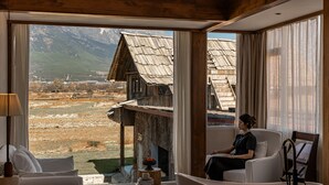 Una habitación con un ventanal amplio que ofrece vistas a edificaciones rústicas y montañas. Hay una silla blanca y una mesa de madera con un objeto decorativo.
