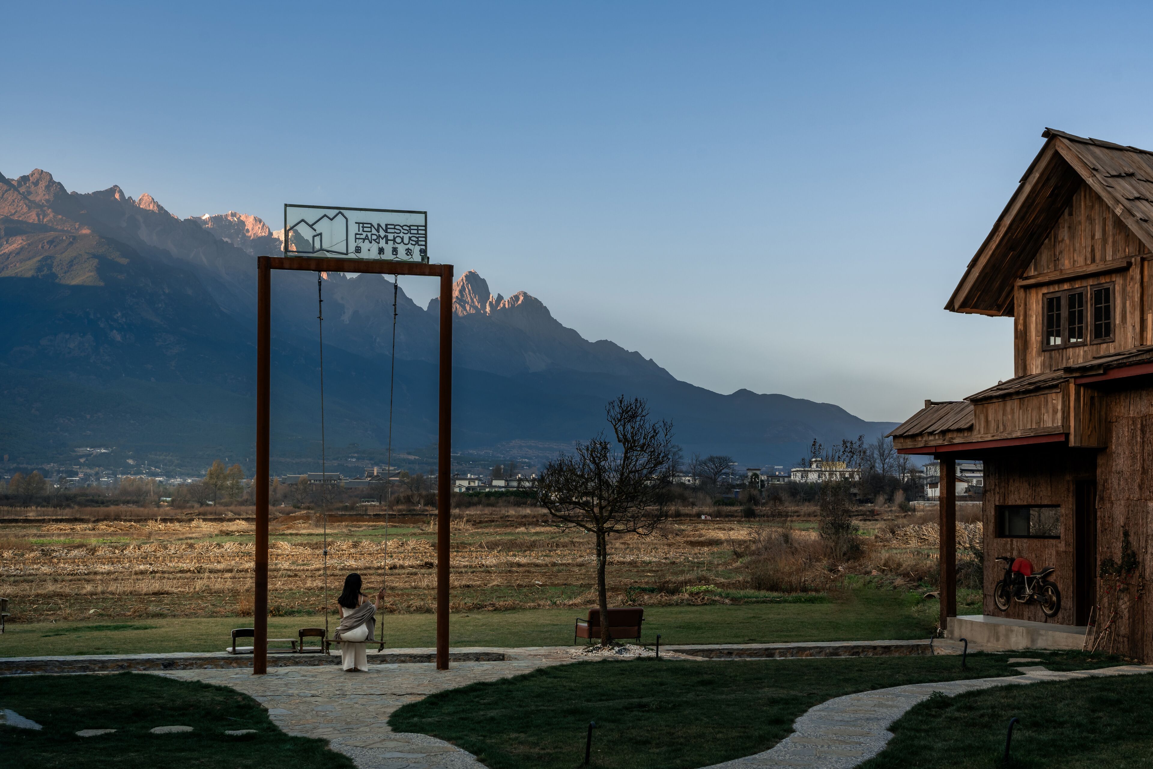 A wooden house with a mountain backdrop, a signboard, and a paved path leading to a bench.