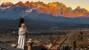 Eine Frau in einem weißen Kleid blickt auf eine gebirgige Landschaft mit Blick ins Tal und entfernte Gebäude.