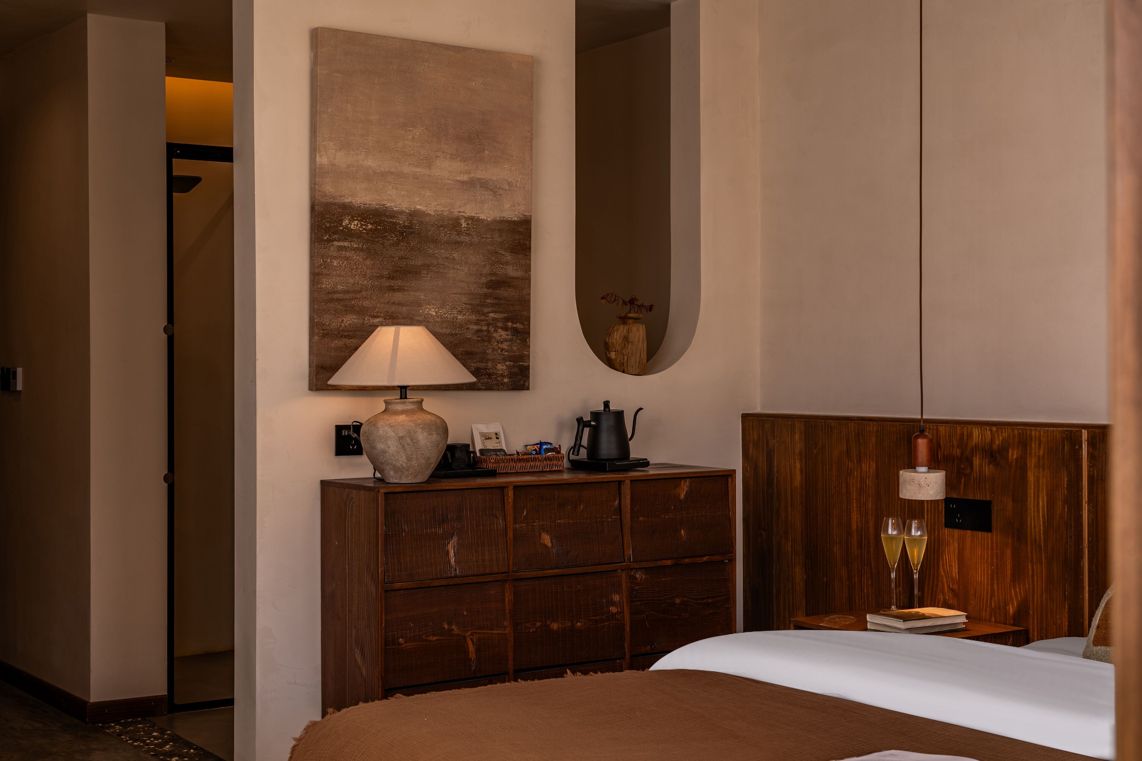 Een hotelkamer met een bed, een houten dressoir, een lamp, een vaas met bloemen en twee glazen wijn op een nachtkastje.
