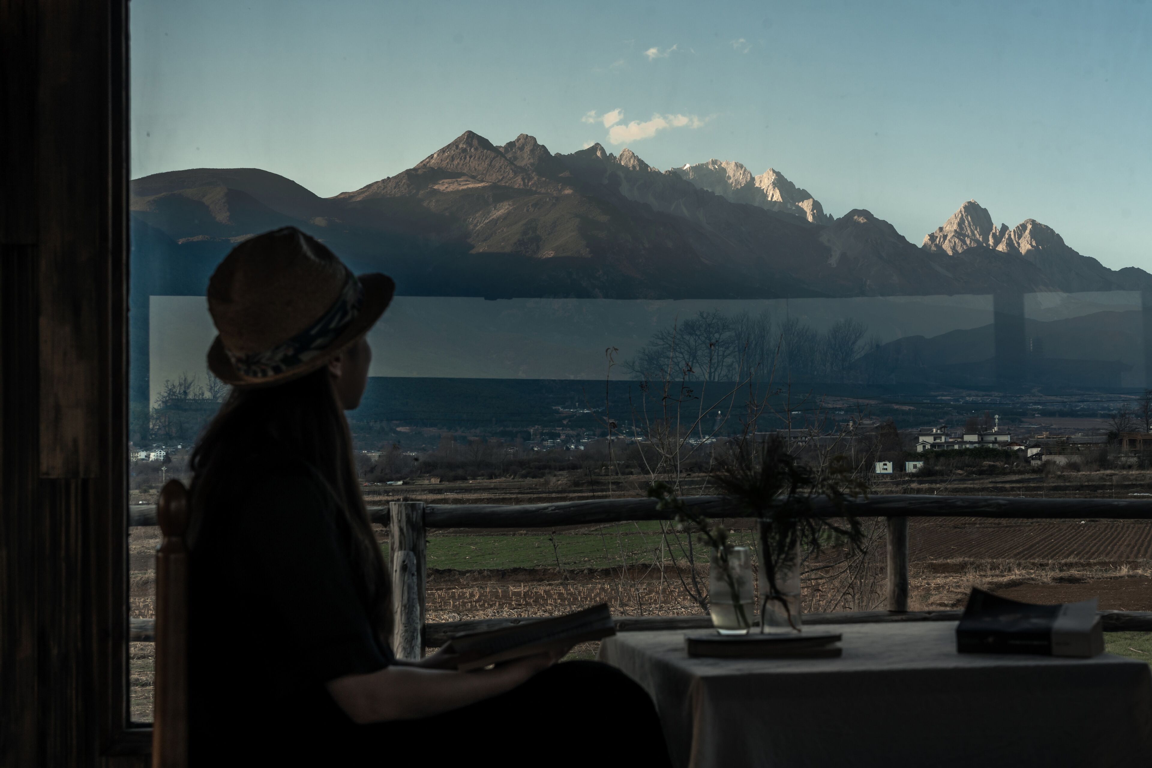 A person seated at a table with a view of mountains and a bridge in the distance.