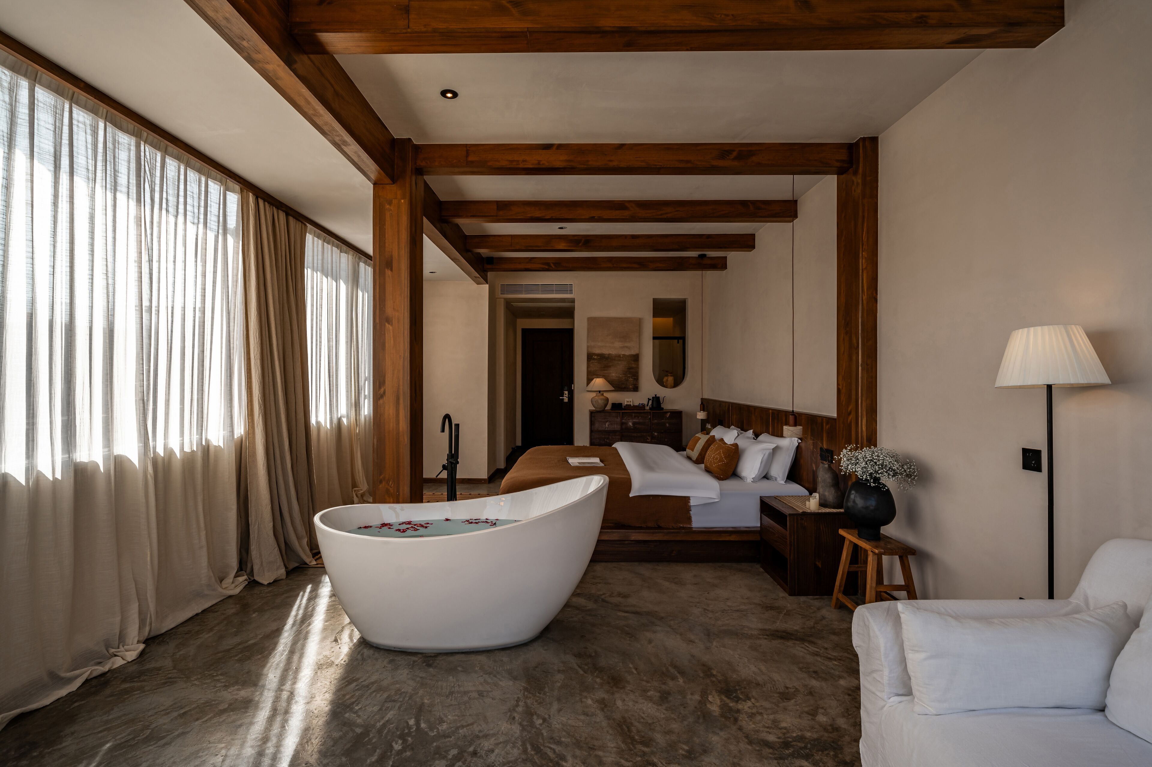 A modern hotel room with a large bathtub, a bed with a wooden headboard, and a white sofa.