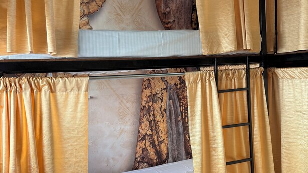 Una cama litera con cortinas amarillas, una escalera de madera, y una pared con una estatua de Buda y motivos florales.