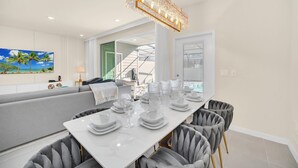 Een moderne eetruimte met een witte tafel voor zes personen, grijs gestoffeerde stoelen en een kristallen kroonluchter.