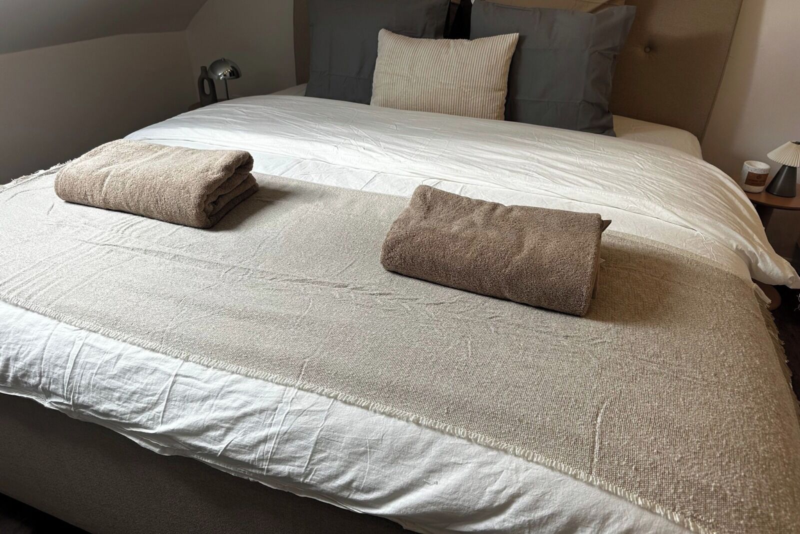 Una cama bien hecha con colcha blanca y color beige, dos toallas dobladas y un cabecero con almohadas.