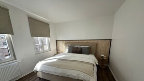 Ein Schlafzimmer mit Bett, Fenster mit Jalousie, Nachttisch mit Lampe und Heizkörper.