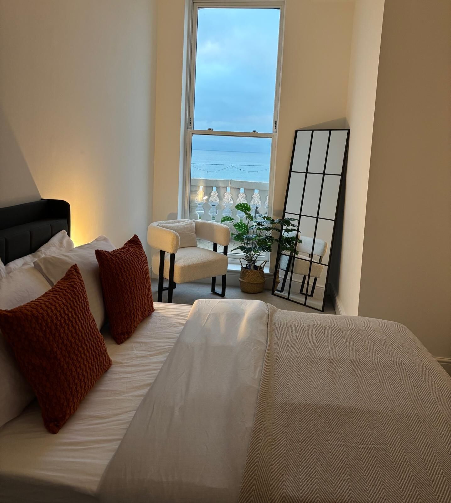 A bedroom with a bed, two chairs, a potted plant, and a view of the sea through a window.