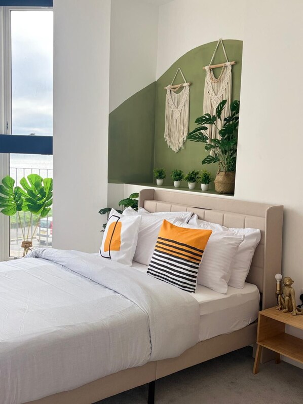A modern bedroom with a bed, macramé wall hangings, potted plants, and a view of the outdoors. - Luxury 2 Bed Apt Seaview By Urban Roost (Worthing)
