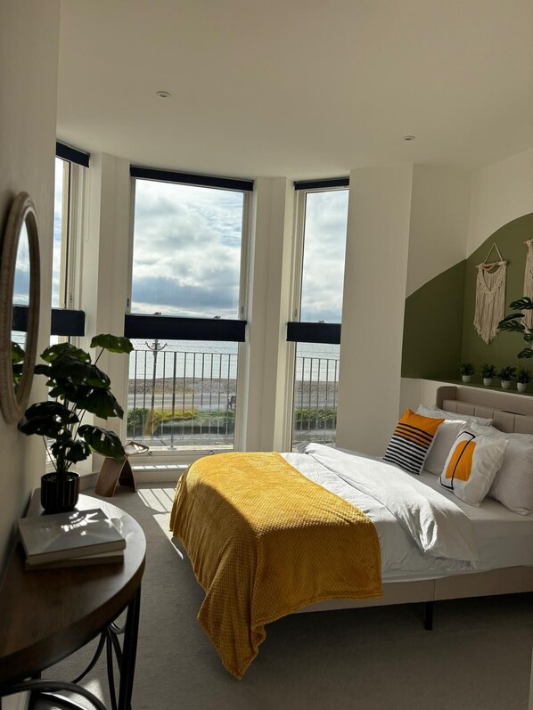 Un dormitorio con cama, escritorio, planta en maceta y vistas al mar a través de amplias ventanas.