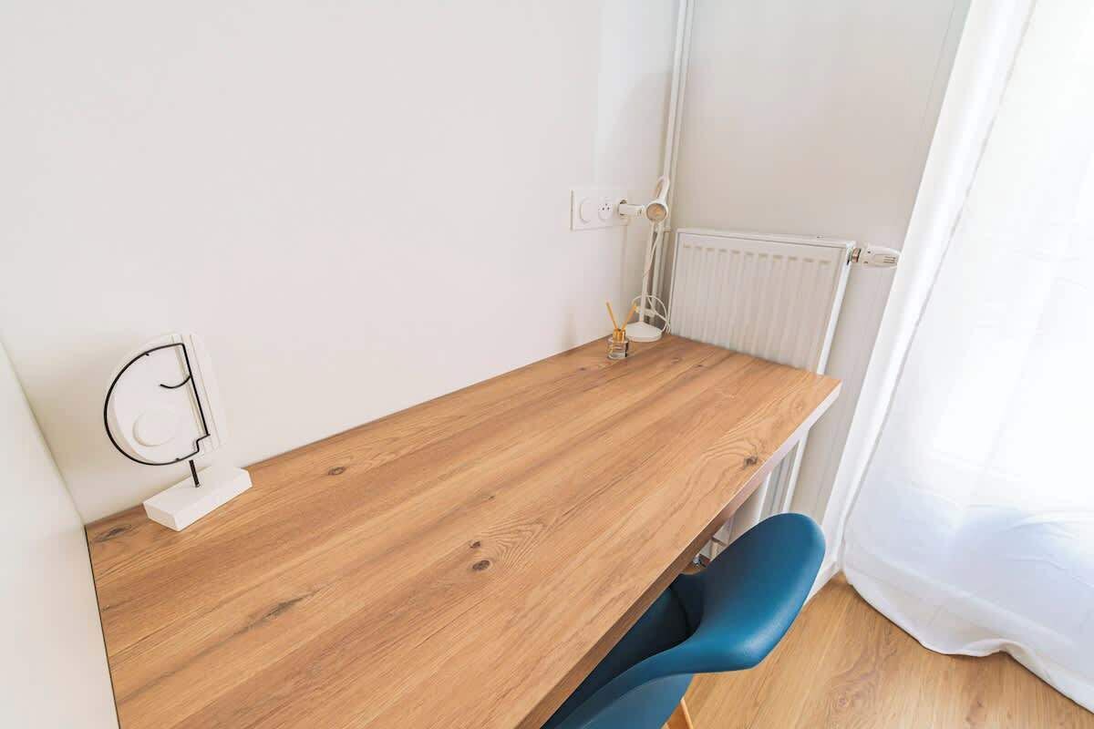 Una habitación pequeña con un escritorio de madera, una silla azul, un radiador blanco y una cortina blanca.