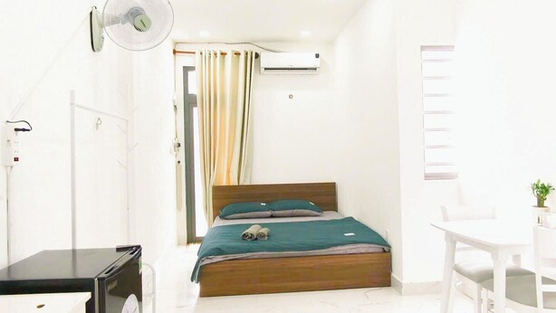 Une chambre d’hôtel de taille réduite, comprenant un lit, un bureau, une chaise, un petit réfrigérateur et un ventilateur de plafond.